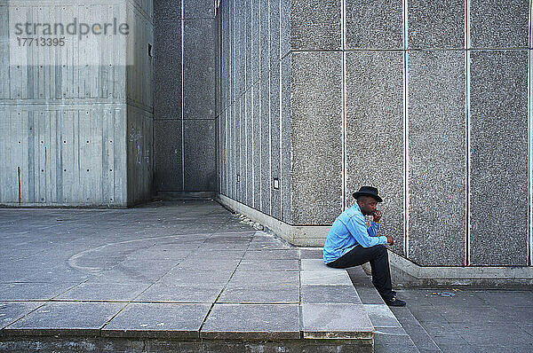 Ein Mann mit blauem Hemd und Hut sitzt auf einer Stufe neben einer Betonmauer  Southbank; London  England