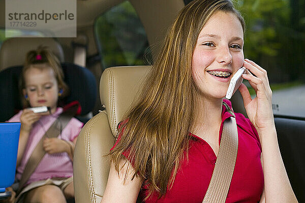 Teenager  der lächelt  während er ein Mobiltelefon benutzt  mit Geschwistern im Hintergrund  die mit Elektronik in einem Mini-Van spielen  Bradford  Ontario