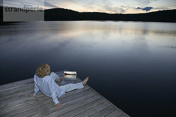 Frau  die einen Laptop auf einem Cottage Dock benutzt  während sie den Sonnenuntergang beobachtet  Ontario