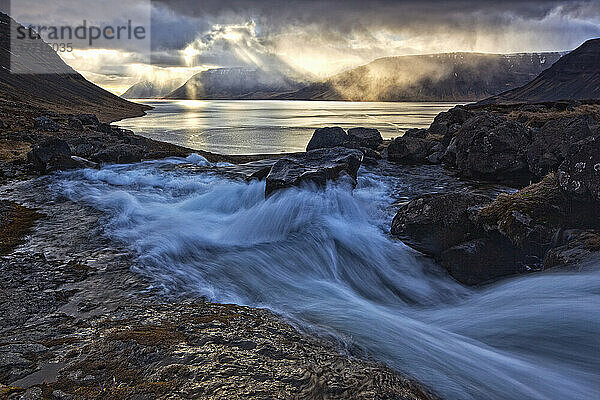 Ein stürmischer Himmel bricht über dem Arnarfjorour-Fjord bei Sonnenuntergang auf  als der Wasserfall Dynjandi in ihn hineinfließt; Island
