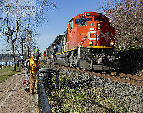 Eine junge Familie steht auf einem Wanderweg an der Küste von British Columbia und beobachtet  wie ein Güterzug auf den Gleisen vorbeifährt; White Rock  British Columbia  Kanada