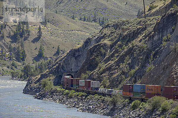 Ein Transportzug mit bunten Schiffscontainern fährt auf den Gleisen neben dem Fraser River im Fraser Canyon und durch einen Tunnel im Berg; British Columbia  Kanada