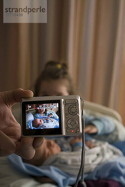 Große Schwester hält neugeborenen Bruder im Krankenhaus  durch die Kamera.