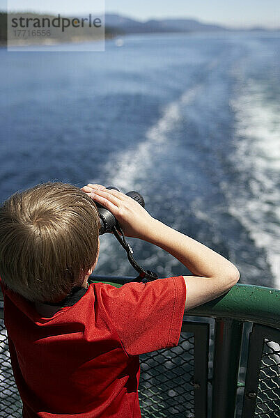 Junge auf dem Achterdeck einer Fähre  der mit einem Fernglas in die Ferne blickt  Fähren-Wake sichtbar.