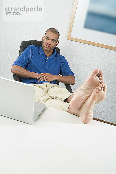 Junger Mann mit Füßen auf dem Schreibtisch mit Blick auf den Computer