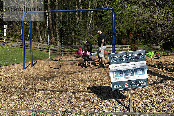 Eine junge Familie spielt auf einem Spielplatz in der Nachbarschaft. Im Vordergrund steht ein Schild  das die Gemeinschaft daran erinnert  sicher und gesund zu bleiben und Abstand zu halten; North Vancouver  British Columbia  Kanada