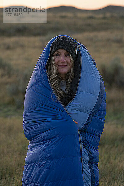 Eine Frau wickelt sich bei Sonnenaufgang in einen Schlafsack ein  um sich warm zu halten  während sie in der weiten Prärielandschaft steht; Val Marie  Saskatchewan  Kanada
