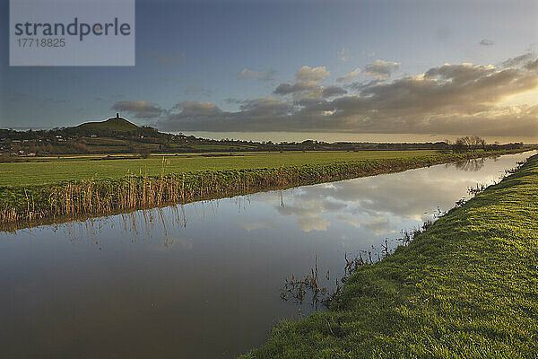 Das ruhige Wasser des Flusses Brue  in dem sich der Himmel spiegelt und das durch die Landschaft in der Nähe von Glastonbury fließt  mit Glastonbury Tor im Hintergrund  Somerset  Großbritannien; Somerset  England