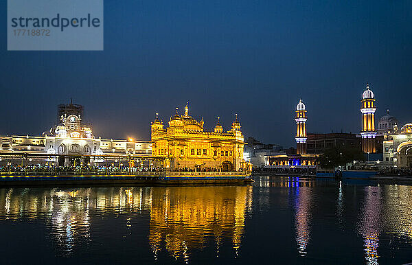 Der Uhrenturm und der Eingang im gotischen Stil zum vergoldeten Goldenen Tempel (Harmandir Sahib)  dem bedeutendsten heiligen Gurdwara-Komplex der Sikh-Religion  mit dem Sarovar (Heiliger Teich) und den Türmen von Ramgarhia Bunga  die bei Nacht beleuchtet sind; Amritsar  Punjab  Indien