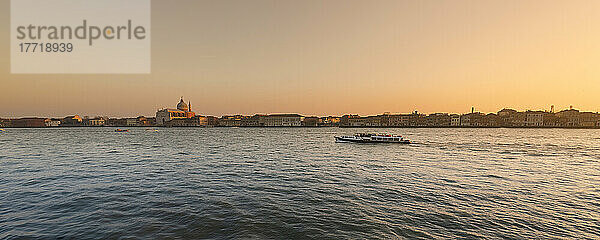 Glühender Sonnenuntergang über der Insel Giudecca  während ein Boot die ruhige venezianische Lagune hinunterfährt und die Kirche Il Redentore in der Skyline zu sehen ist; Giudecca  Italien
