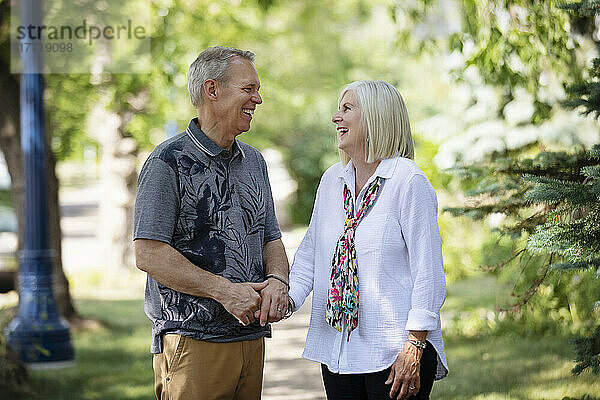 Älteres Paar  das sich an den Händen hält und einen Moment gemeinsam lacht  während es im Freien auf einem Parkweg spazieren geht; Edmonton  Alberta  Kanada