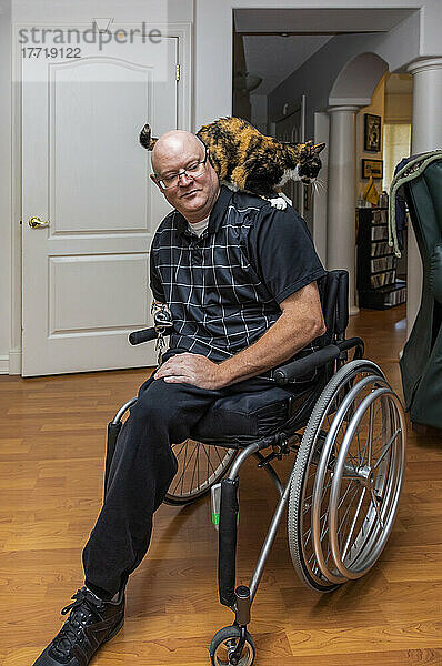 Mann mit doppelter Gliedmaßenamputation  der in seinem Haus sitzt  während seine Hauskatze über seine Schultern krabbelt; St. Albert  Alberta  Kanada