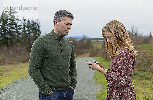 Paar im mittleren Erwachsenenalter steht in einem Park  die Frau benutzt ein Smartphone  während ihr Mann sie beobachtet; Aldergrove  British Columbia  Kanada