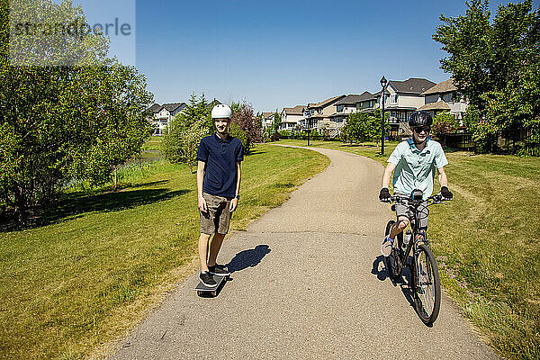 Brüder  die gemeinsam Zeit in der Nachbarschaft verbringen  einer auf einem Fahrrad und einer auf einem Skateboard; Edmonton  Alberta  Kanada