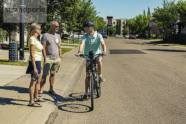 Junger Mann  der mit seinem Fahrrad eine Wohnstraße hinunterfährt und anhält  um mit seinen Eltern zu sprechen  die auf dem Bürgersteig stehen; Edmonton  Alberta  Kanada