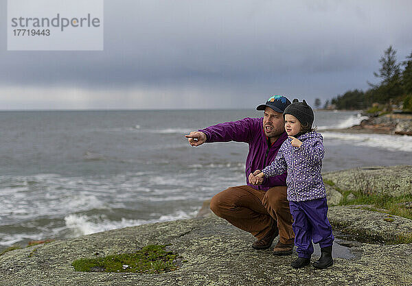 Ein Vater hält die Hand seiner kleinen Tochter  während sie am Ufer stehen und auf das raue Wasser des Ozeans hinausschauen  Sunshine Coast  BC  Kanada; British Columbia  Kanada