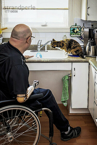 Mann mit doppelter Gliedmaßenamputation arbeitet zu Hause in der Küche  während seine Hauskatze Wasser aus dem Wasserhahn des Spülbeckens trinkt; St. Albert  Alberta  Kanada