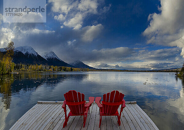 Zwei rote Stühle auf einer Terrasse am ruhigen Lake Kathlyn  umgeben von herbstlich gefärbten Bäumen und schneebedeckten Coast Mountains  vom Lakedrop Inn aus gesehen  Watson's Landing  BC  Kanada; Smithers  British Columbia  Kanada
