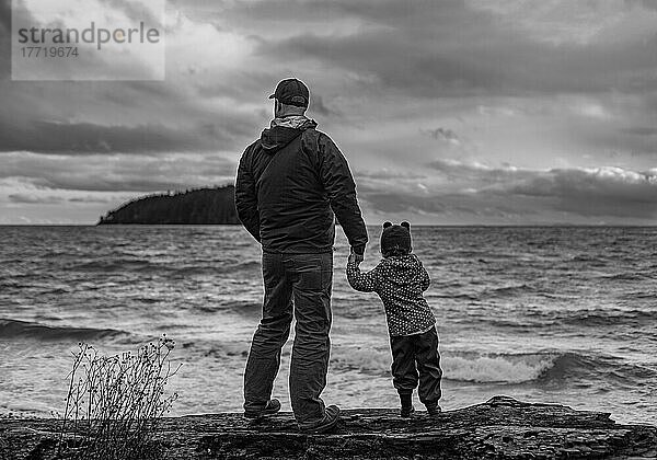 Ein Vater hält die Hand seiner kleinen Tochter  während sie am Ufer stehen und auf das raue Wasser des Ozeans hinausschauen  Sunshine Coast  BC  Kanada; British Columbia  Kanada