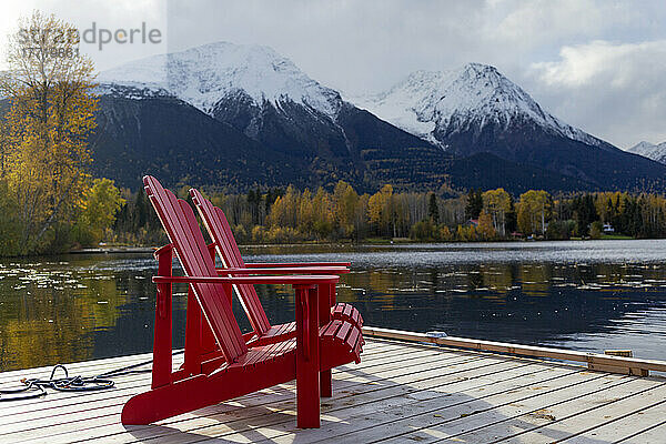 Zwei rote Stühle auf einer Terrasse am ruhigen Lake Kathlyn  umgeben von herbstlich gefärbten Bäumen und schneebedeckten Coast Mountains  vom Lakedrop Inn aus gesehen  Watson's Landing  BC  Kanada; Smithers  British Columbia  Kanada