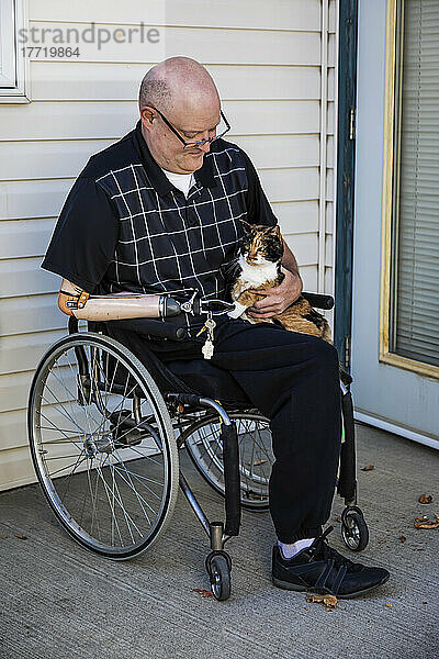 Mann mit doppelter Gliedmaßenamputation sitzt vor seinem Haus mit seiner Hauskatze auf dem Schoß; St. Albert  Alberta  Kanada