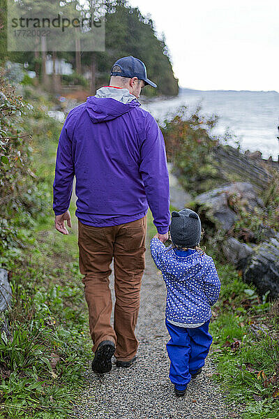 Ein Vater hält die Hand seiner kleinen Tochter  während sie auf einem Weg zum Strand an der Sunshine Coast  BC  Kanada  spazieren gehen; British Columbia  Kanada