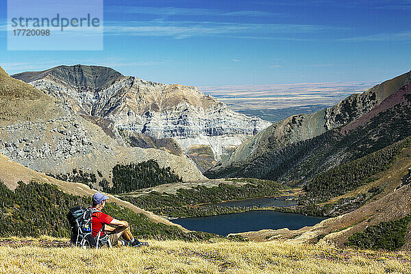 Männlicher Wanderer  der sich auf einem grasbewachsenen Bergrücken ausruht  mit Blick auf einen Alpensee  Bergketten  blauer Himmel und Wolken im Hintergrund  Waterton Lakes National Park; Waterton  Alberta  Kanada