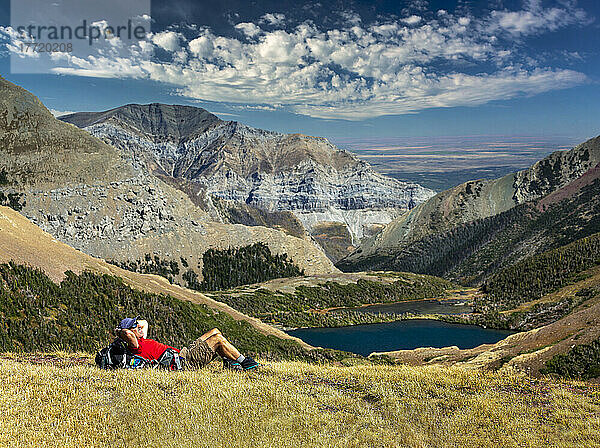 Männlicher Wanderer  der sich auf einem grasbewachsenen Bergrücken ausruht  mit Blick auf einen Alpensee  Bergketten  blauer Himmel und Wolken im Hintergrund  Waterton Lakes National Park; Alberta  Kanada