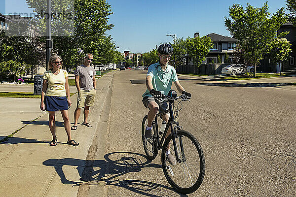 Junger Mann fährt mit seinem Fahrrad eine Wohnstraße hinunter  seine Eltern stehen auf dem Bürgersteig und schauen zu; Edmonton  Alberta  Kanada