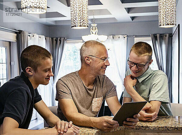 Vater und zwei Söhne sitzen zu Hause an der Kücheninsel  benutzen ein Tablet und reden miteinander; Edmonton  Alberta  Kanada