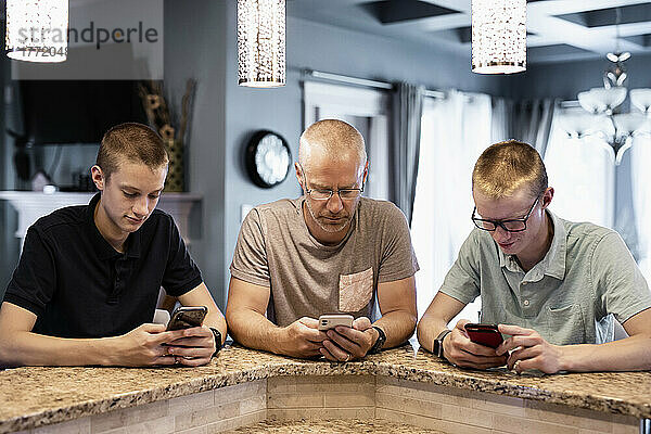 Vater und zwei Söhne sitzen an der Kücheninsel zu Hause und benutzen ihre Smartphones; Edmonton  Alberta  Kanada