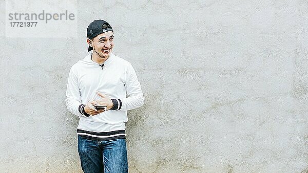 Ein junger lateinamerikanischer Mann lacht mit seinem Handy  das an einer Wand lehnt  ein gutaussehender Mann  der mit seinem Handy glücklich ist  ein junger lateinamerikanischer Mann  der mit seinem Handy glücklich ist und an einer Wand lehnt
