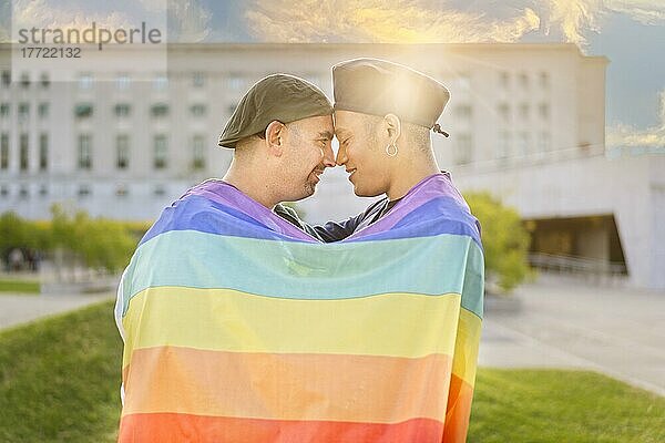 Verliebtes schwules Paar  das sich gegenseitig umarmt  eingewickelt in eine schwule Flagge  in einem Park bei Sonnenuntergang  mit Reflexionen der Sonne. Konzept der Vielfalt  Stolz  Liebe  Gleichheit