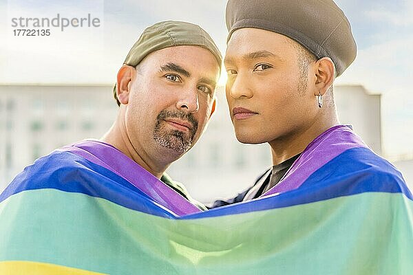 Verliebtes schwules Paar  das sich gegenseitig umarmt  eingewickelt in eine schwule Flagge  in einem Park bei Sonnenuntergang  mit Reflexionen der Sonne. Konzept der Vielfalt  Stolz  Liebe  Gleichheit