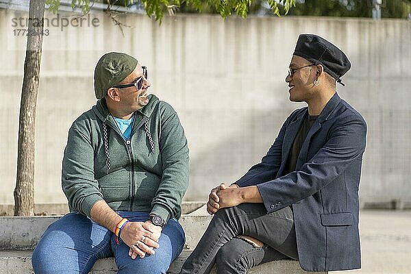 Homosexuelles lateinamerikanisches Paar  das auf einer Bank in einem Park sitzt  modische Hüte und Sonnenbrillen trägt und sich lächelnd ansieht