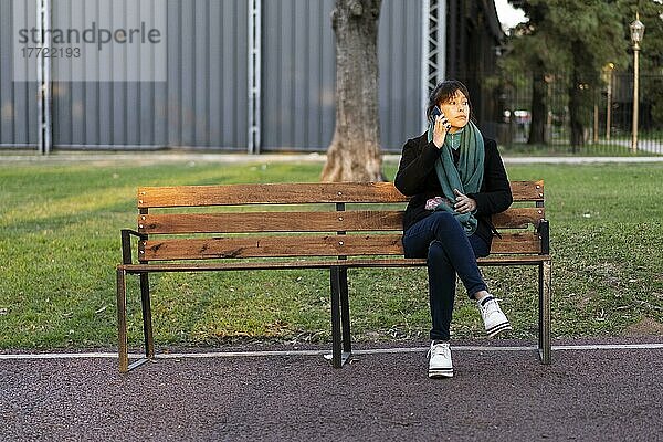 Lateinamerikanische Frau  die auf einer Bank im Park sitzt und mit einem Handy telefoniert