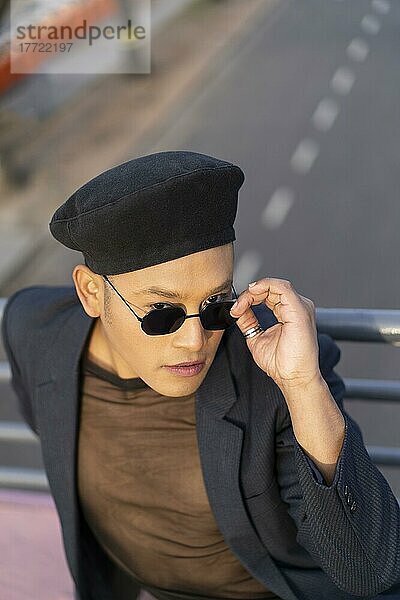 Latino schwuler Mann mit Make-up und modischem Hut posiert auf einer Brücke