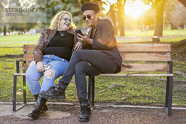 Schwule Freunde sitzen bei Sonnenuntergang auf einer Bank im Park  unterhalten sich und schauen auf ihr Handy