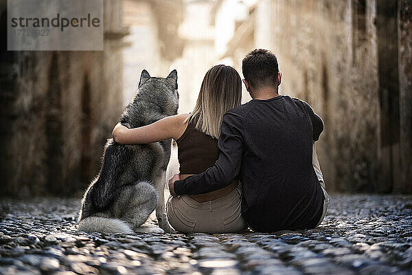 Rückansicht von zwei jungen Menschen und ihrem geliebten Wolfshund  die in einer Altstadtstraße sitzen und sich umarmen und in die Sonne starren  Piemont  Italien  Europa