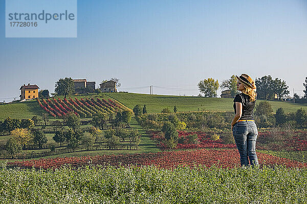 Junge Frau mit blonden Haaren und Hut blickt auf einen Hügel  der im Herbst von roten Weinbergen bedeckt ist  Emilia Romagna  Italien  Europa