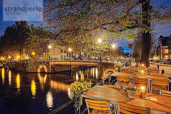 Nachtansicht von Amsterdam mit Cafétischen  Gracht  Brücke und mittelalterlichen Häusern in der Abenddämmerung beleuchtet. Amsterdam  Niederlande  Europa