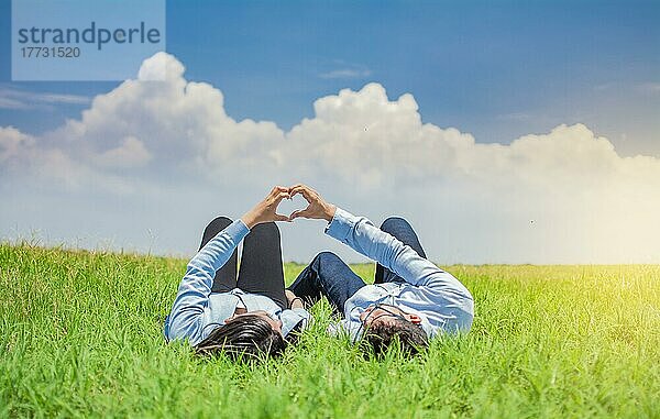 Happy couple lying on graß making heart shape  Couple on graß making heart shape with hands  Young couple lying on graß making love symbol with hands