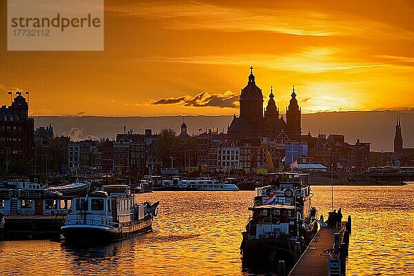 Amsterdamer Stadtsilhouette mit der Sankt-Nikolaus Kirche (Sint-Nicolaaskerk) und Booten auf dem Pier bei Sonnenuntergang mit dramatischem Himmel. Amsterdam  Niederlande  Europa