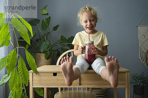 Happy girl sitting on table holding smoothie at home