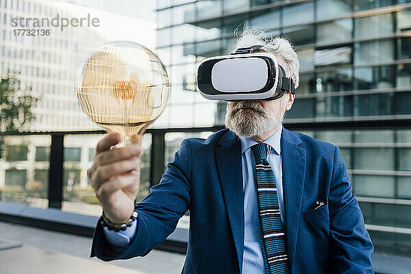 Leitender Geschäftsmann hält Glühbirne und trägt Virtual-Reality-Simulator auf dem Bürobalkon