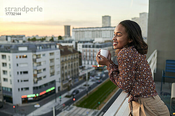Lächelnde junge Frau hält bei Sonnenuntergang eine Kaffeetasse auf dem Balkon
