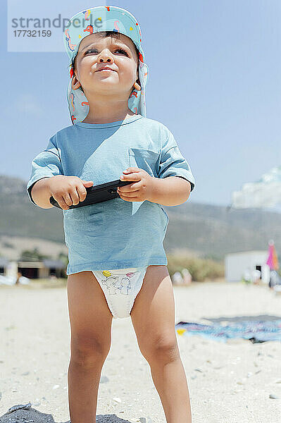 Junge mit Mobiltelefon am Strand an einem sonnigen Tag