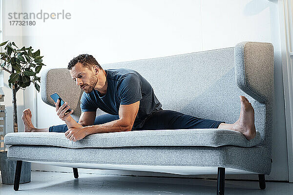 Mann sitzt mit gespreizten Beinen zu Hause auf dem Sofa und benutzt ein Smartphone