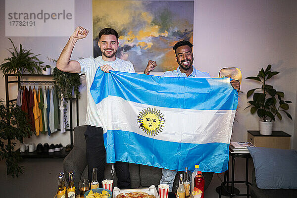 Cheerful young men gesturing fists standing with Argentinian flag at home