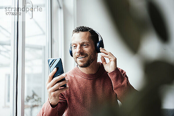 Lächelnder Mann hört Musik über Kopfhörer und hält zu Hause sein Smartphone in der Hand